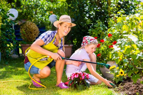In a lush, well-maintained garden, a woman wearing a straw hat, yellow apron, and casual clothing is kneeling on a neatly trimmed green lawn, assisting a young girl in planting flowers. The girl, dressed in a light-coloured outfit and a floral sun hat, is spreading soil around vibrant pink and red blossoms in a flower bed bordered by dark soil. The garden features a variety of flowering plants, including white and yellow blooms, with dense green foliage and tall bushes in the background. Bright sunlight illuminates the scene, highlighting the natural textures of the grass, soil, and petals, creating a cheerful outdoor environment suitable for gardening services in Balham. A garden hose with a spray nozzle is held by the woman, suggesting active maintenance, and the surrounding area includes potted plants and a garden fence covered with leafy vines, contributing to a local, well-established garden setting typical of residential outdoor spaces in the Balham area. This scene underscores professional gardening and lawn care activities provided by Hedge Trimming Balham, with a focus on cultivating vibrant flowerbeds and maintaining a healthy garden environment.